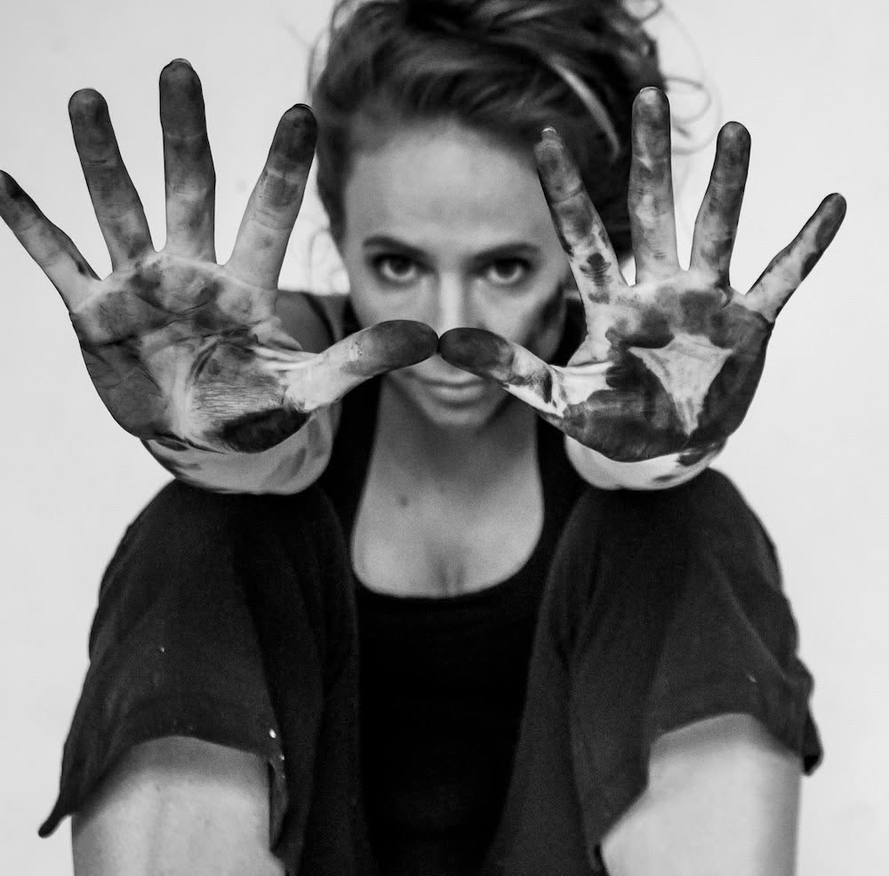 Black and white portrait of Kylian holding both paint-covered palms up to the camera, fingertips touching just below the eyes.